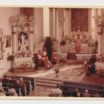 Priester in roten Gewändern zelebrieren einen feierlichen Gottesdienst in einer barocken Kirche vor Gläubigen.