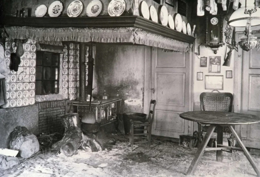 Schwarz-Weiß-Foto einer verwüsteten Bauernstube mit Kachelherd, Petroleumlampe und Dekortellern an der Decke.
