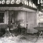 Schwarz-Weiß-Foto einer verwüsteten Bauernstube mit Kachelherd, Petroleumlampe und Dekortellern an der Decke.