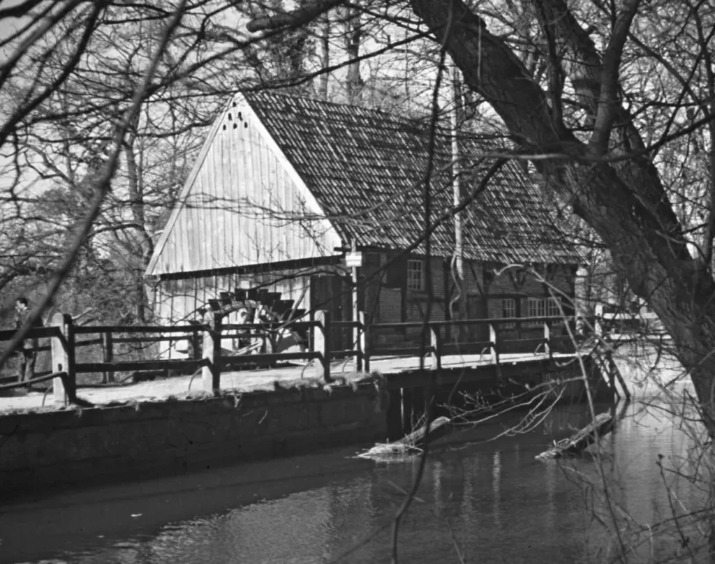 Schwarzweißfoto einer alten Wassermühle mit Mühlrad an einem Fluss, umgeben von kahlen Bäumen.