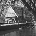 Schwarzweißfoto einer alten Wassermühle mit Mühlrad an einem Fluss, umgeben von kahlen Bäumen.