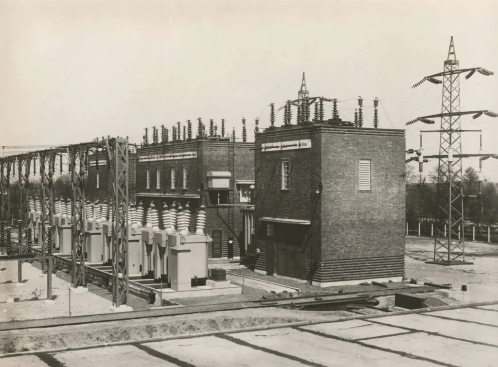 Schwarz-Weiß-Foto eines historischen Elektrizitätswerks mit Backsteingebäuden, Hochspannungsmasten und Isolatoren.