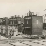 Schwarz-Weiß-Foto eines historischen Elektrizitätswerks mit Backsteingebäuden, Hochspannungsmasten und Isolatoren.