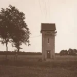 Kleiner gemauerter Transformatorturm mit Ziegeldach auf einer Wiese, mit Stromleitungen verbunden, um 1920.