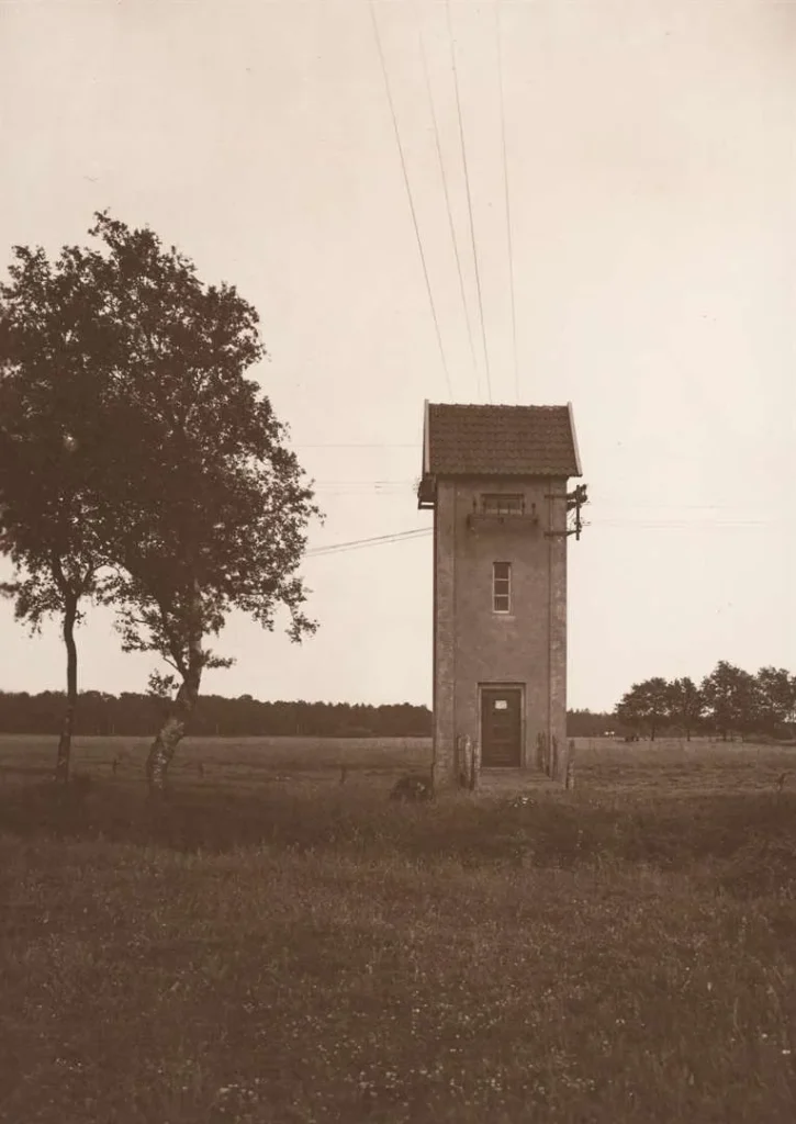 Kleiner gemauerter Transformatorturm mit Ziegeldach auf einer Wiese, mit Stromleitungen verbunden, um 1920.