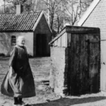 Eine ältere Frau steht neben einem gemauerten Brunnen mit Holzabdeckung auf einem Bauernhof, um 1950.