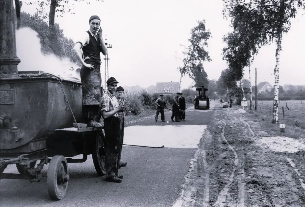 Arbeiter beim Teerieren einer Landstraße im Emsland, mit dampfendem Teerkocher und Straßenwalze im Hintergrund.