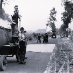 Arbeiter beim Teerieren einer Landstraße im Emsland, mit dampfendem Teerkocher und Straßenwalze im Hintergrund.