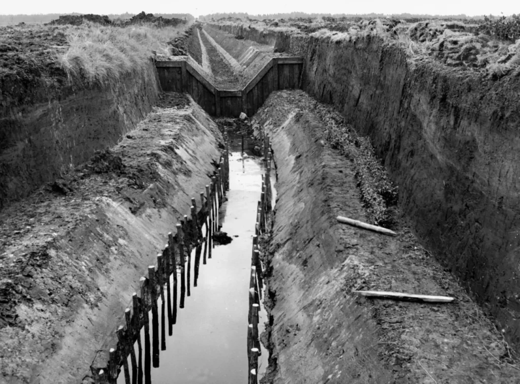 Tiefer Erdaushub eines kanalisierten Flusslaufs mit Holzpfählen und Stauwehr im Emsland.