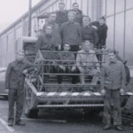 Gruppe junger Männer in Arbeitskleidung posiert auf und neben einem Mähdrescher vor einem Industriegebäude.