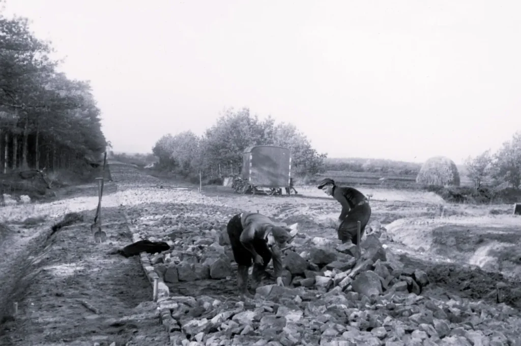 Zwei Arbeiter verlegen Pflastersteine auf einer Baustelle im Emsland, im Hintergrund ein Bauwagen und Bäume.