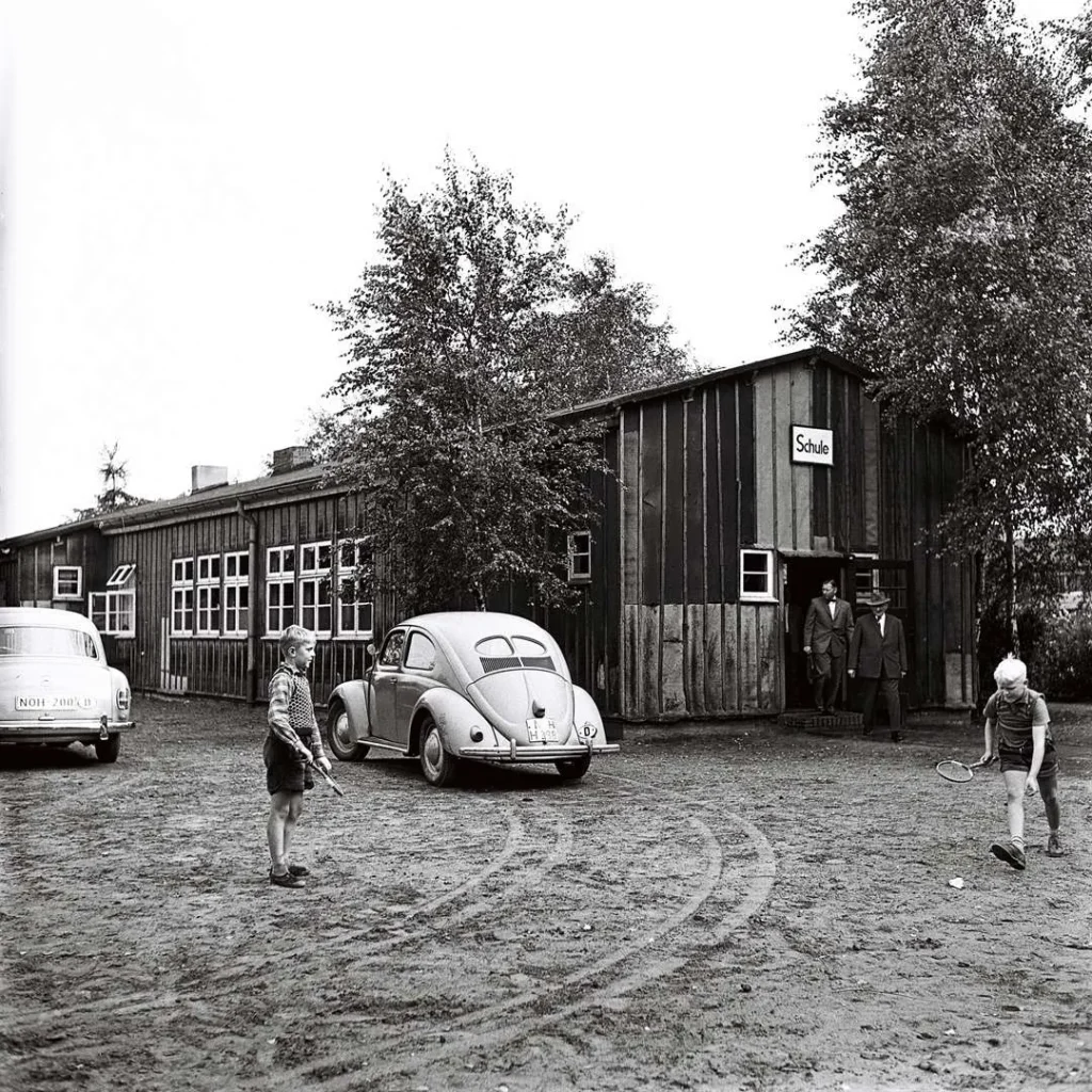 Kinder spielen Federball vor einer Schulbaracke mit Schild 'Schule', daneben ein VW Käfer und zwei Männer im Eingang.