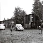Kinder spielen Federball vor einer Schulbaracke mit Schild 'Schule', daneben ein VW Käfer und zwei Männer im Eingang.