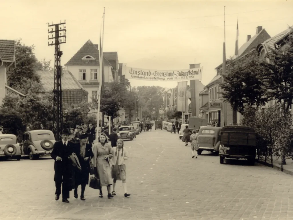 Belebte Straße in Meppen 1951 mit Banner zur Emsland-Ausstellung, Passanten und geparkten Autos der Nachkriegszeit.