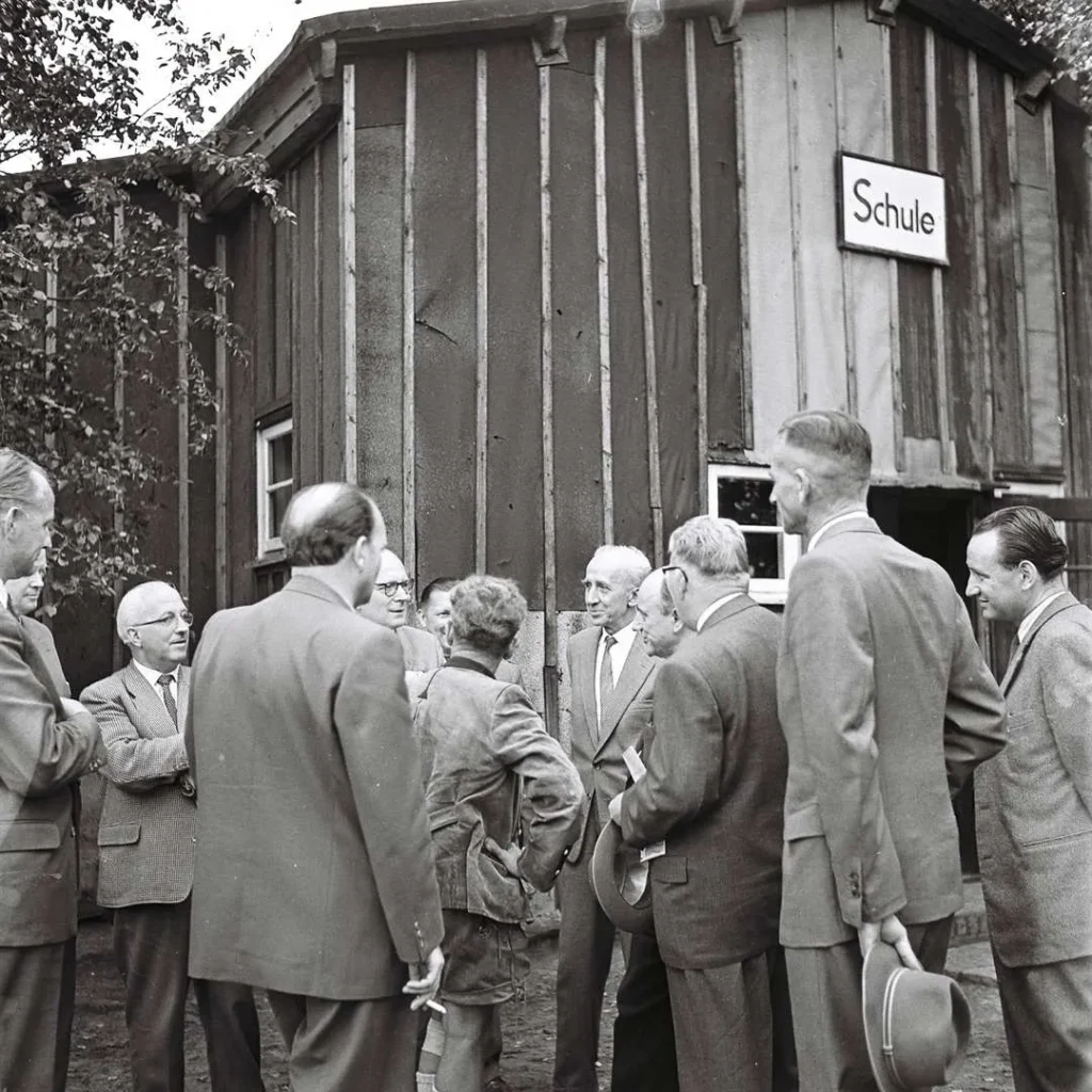 Gruppe von Männern in Anzügen vor einer Holzbaracke mit Schild 'Schule' in Füchtenfeld, August 1958.