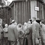 Gruppe von Männern in Anzügen vor einer Holzbaracke mit Schild 'Schule' in Füchtenfeld, August 1958.