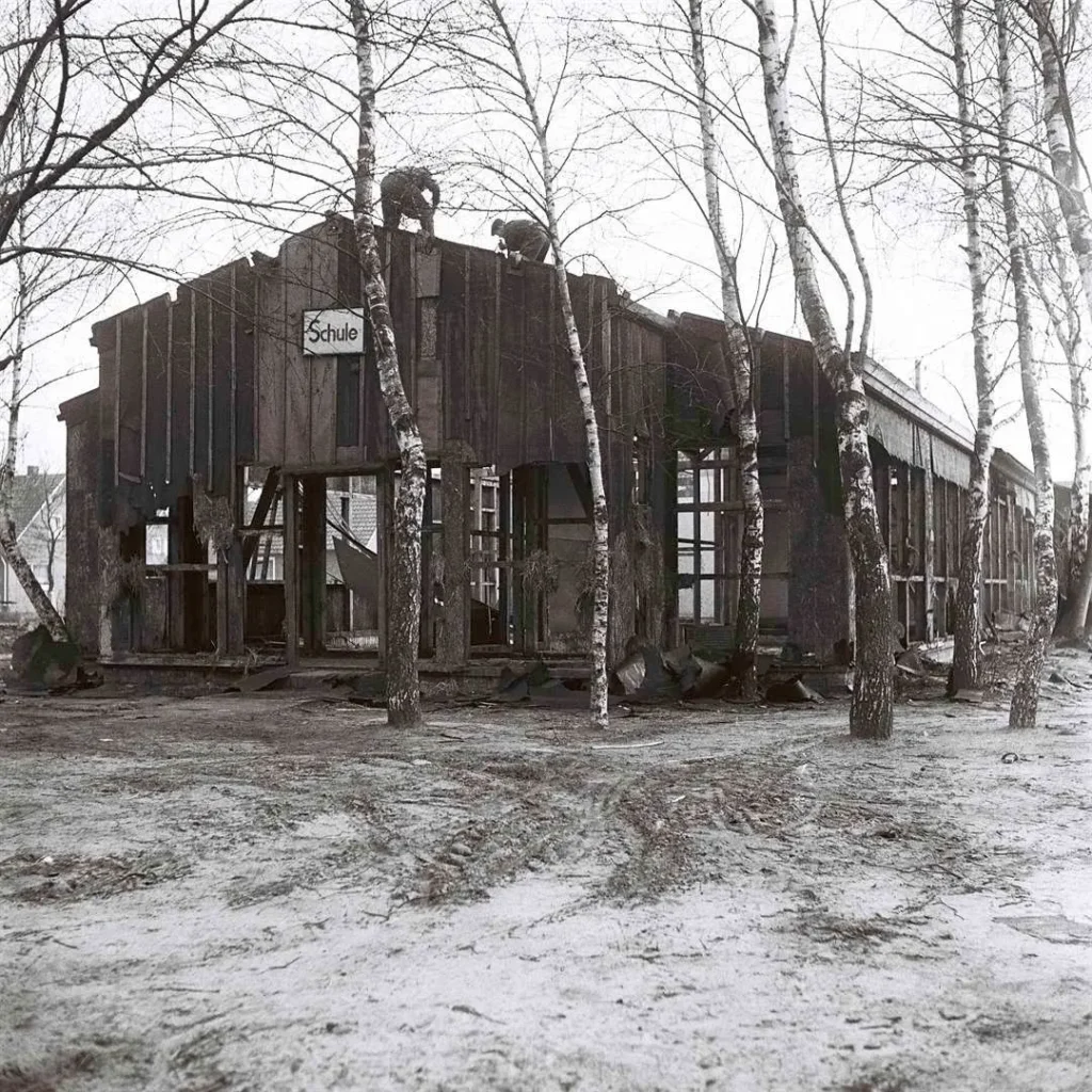 Baufällige Holzbaracke mit Schild 'Schule', umgeben von kahlen Birken; Arbeiter demontieren das marode Dach.