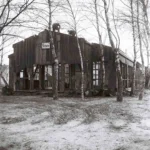 Baufällige Holzbaracke mit Schild 'Schule', umgeben von kahlen Birken; Arbeiter demontieren das marode Dach.