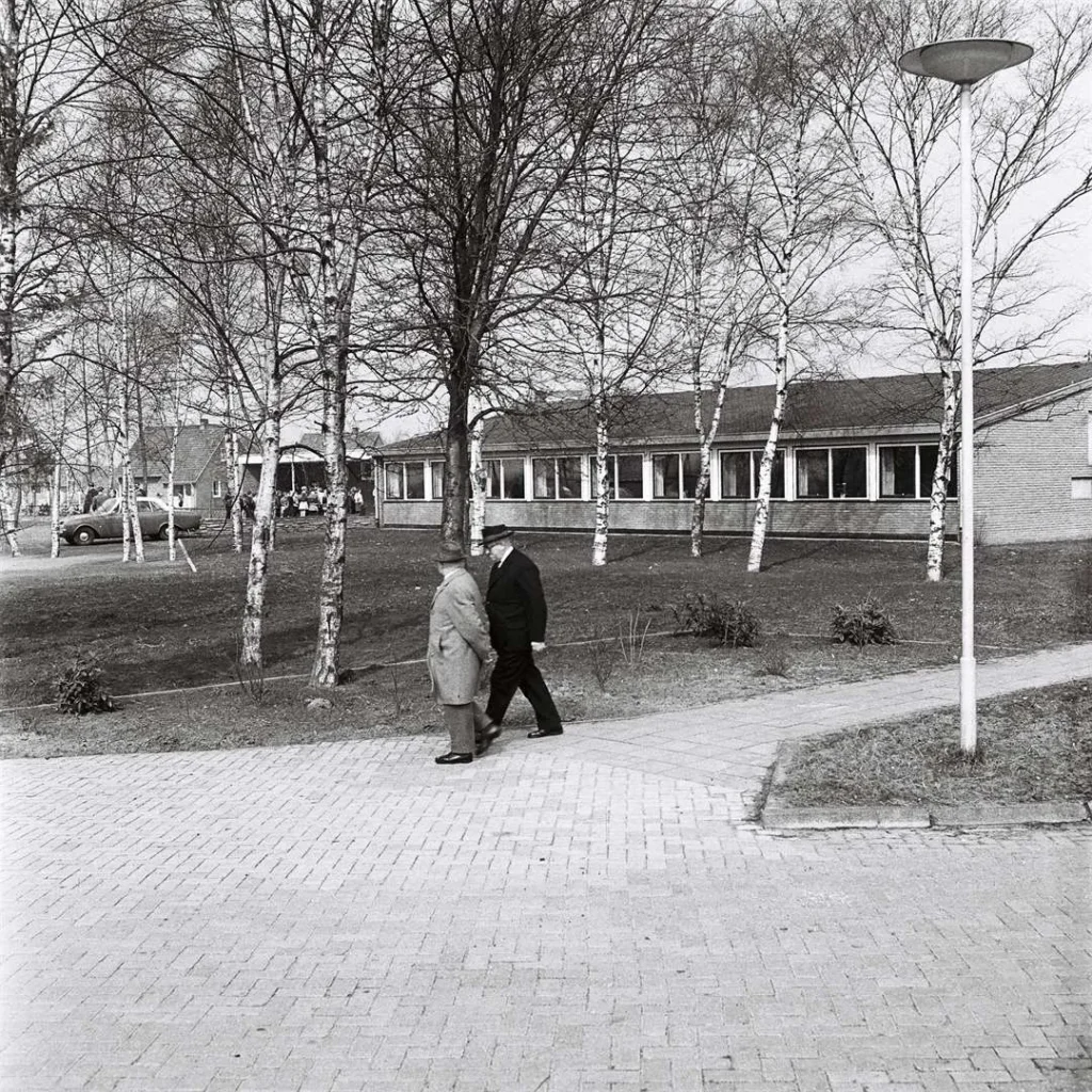 Zwei ältere Männer in Mänteln und Hüten gehen auf einem Gehweg vor einem modernen Flachbau-Schulgebäude spazieren.