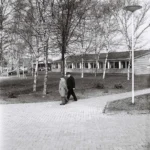 Zwei ältere Männer in Mänteln und Hüten gehen auf einem Gehweg vor einem modernen Flachbau-Schulgebäude spazieren.