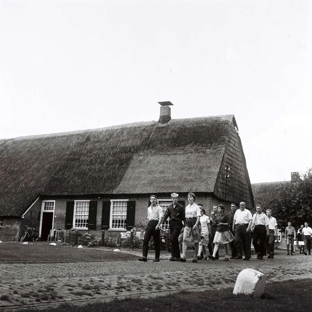 Gruppe von Menschen spaziert auf gepflastertem Weg vor einem reetgedeckten niederländischen Bauernhaus, ca. 1961.