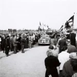 Festumzug zur Brückeneröffnung 1961: Menschenmassen säumen eine Straße, ein Mercedes-Oldtimer fährt durch die Menge.