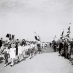 Kinder und Erwachsene marschieren bei der Eröffnung der Grenzbrücke zwischen Emlichheim und Schoonebeek, gesäumt von deutschen und niederländischen Flaggen.