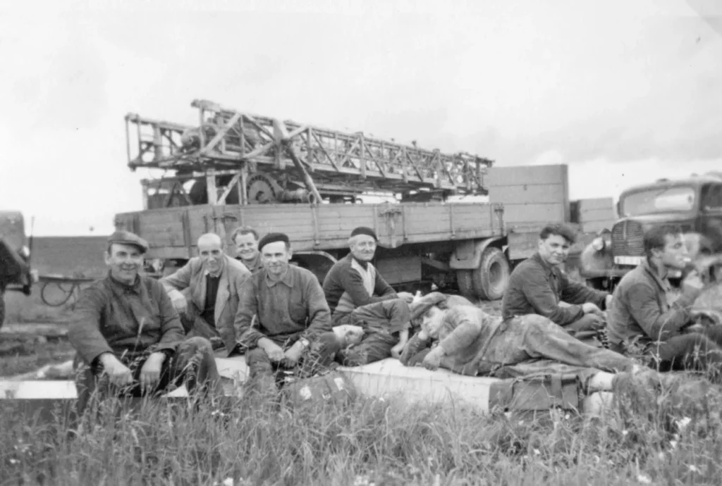 Gruppe von Arbeitern rastet im Gras vor Bohrfahrzeugen auf einem Erdölfeld im Emsland.