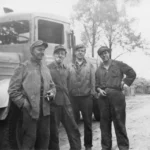 Vier Arbeiter in Arbeitskleidung und Schirmmützen stehen rauchend vor einem LKW auf einem unbefestigten Weg.