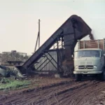 Ein Mercedes-Lkw wird über ein Förderband mit maschinell geworbenem Torf beladen, ein Arbeiter steht daneben.