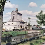 Historische Torfverarbeitungsanlage mit Förderturm an einem Kanal, umgeben von Grünflächen und Wirtschaftsgebäuden.