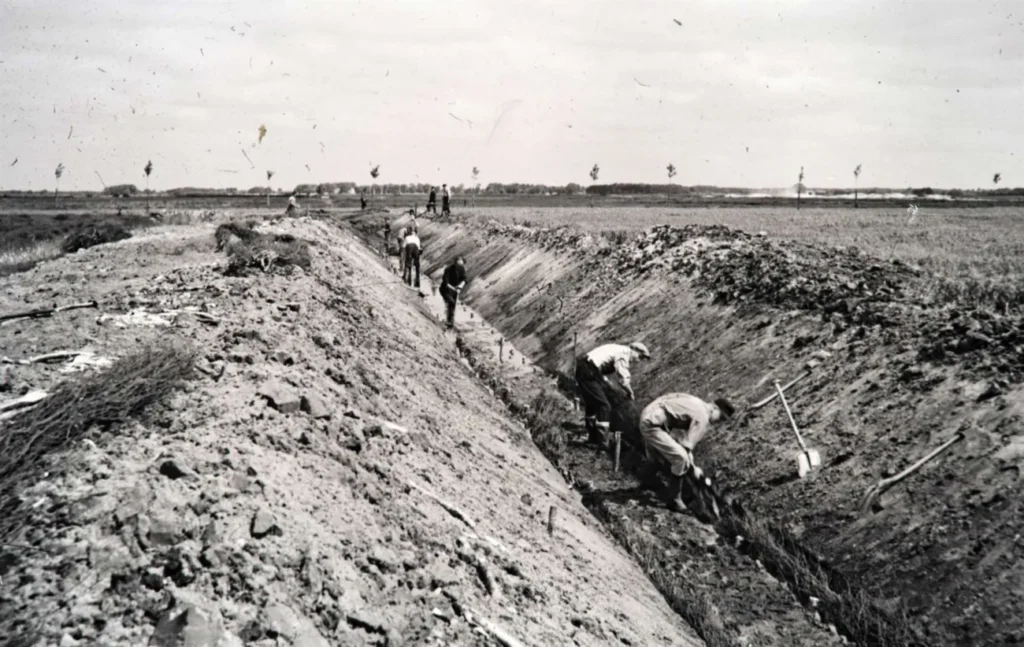 Arbeiter graben manuell einen langen Entwässerungsgraben in flacher Landschaft, aufgenommmen in schwarz-weiß.