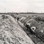 Arbeiter graben manuell einen langen Entwässerungsgraben in flacher Landschaft, aufgenommmen in schwarz-weiß.