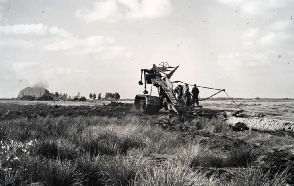Historische Aufnahme einer Gleiskettenbaggermaschine beim Entwässern von Moorland, flankiert von Arbeitern, mit Gehöft im Hintergrund.