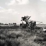 Historische Aufnahme einer Gleiskettenbaggermaschine beim Entwässern von Moorland, flankiert von Arbeitern, mit Gehöft im Hintergrund.