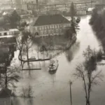 Luftaufnahme einer überfluteten Stadt mit einem Boot zwischen Gebäuden und Bäumen, vermutlich Nordhorn bei Vechte-Hochwasser.