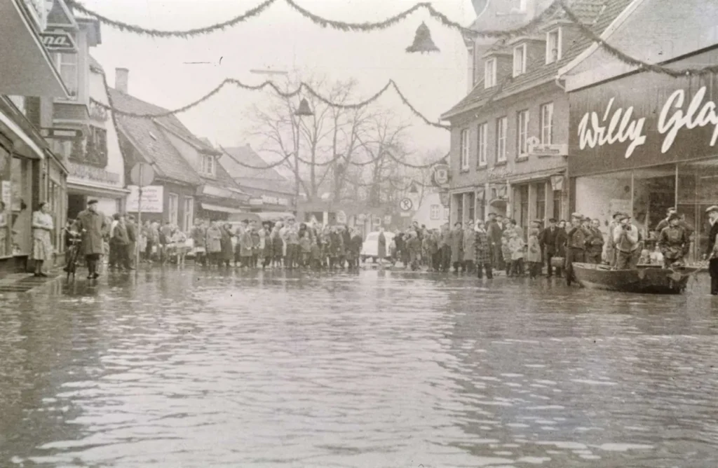 Überflutete Hauptstraße in Nordhorn mit zahlreichen Menschen und einem Boot, im Hintergrund weihnachtliche Straßendekoration.