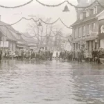 Überflutete Hauptstraße in Nordhorn mit zahlreichen Menschen und einem Boot, im Hintergrund weihnachtliche Straßendekoration.
