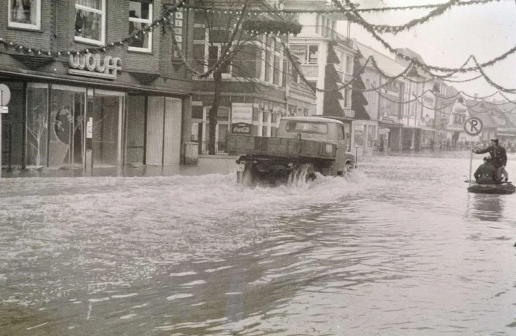 Ein Lastwagen durchfährt eine überflutete Einkaufsstraße in Nordhorn, während ein Mann auf einem Motorrad im Hochwasser sitzt.