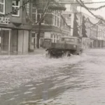 Ein Lastwagen durchfährt eine überflutete Einkaufsstraße in Nordhorn, während ein Mann auf einem Motorrad im Hochwasser sitzt.