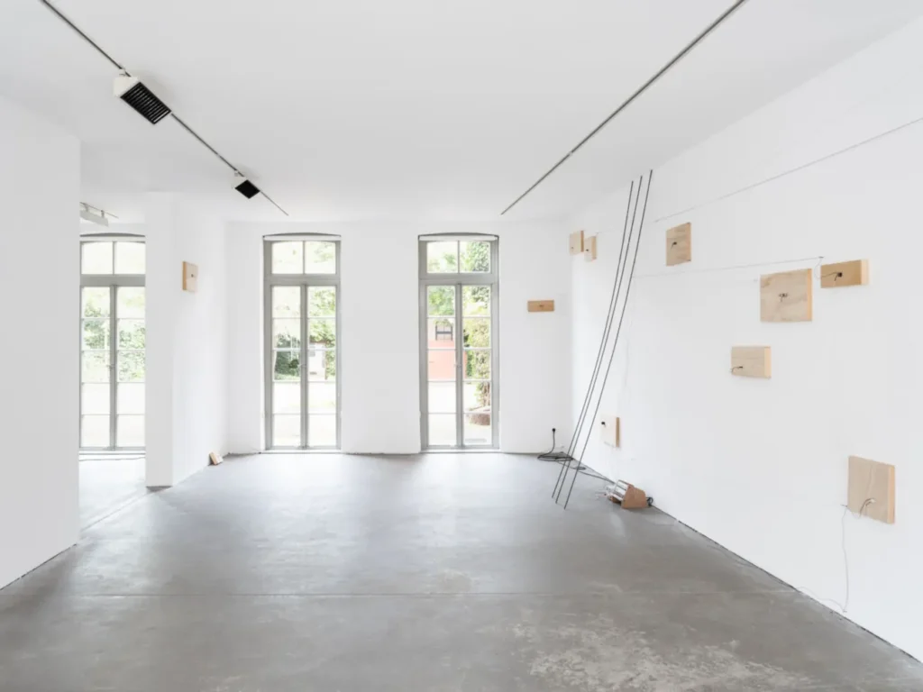 Minimalistische Galerieinstallation mit Holzplatten an weißer Wand, verbunden durch schwarze Drähte, die zum Boden führen.