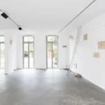 Minimalistische Galerieinstallation mit Holzplatten an weißer Wand, verbunden durch schwarze Drähte, die zum Boden führen.
