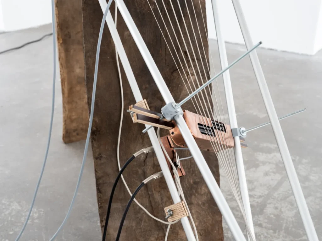 Installationsdetail mit weißen Metallstäben, gespannten Saiten und Holz-Metall-Elementen auf einem Betonsockel.