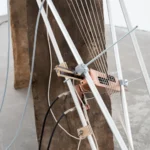 Installationsdetail mit weißen Metallstäben, gespannten Saiten und Holz-Metall-Elementen auf einem Betonsockel.