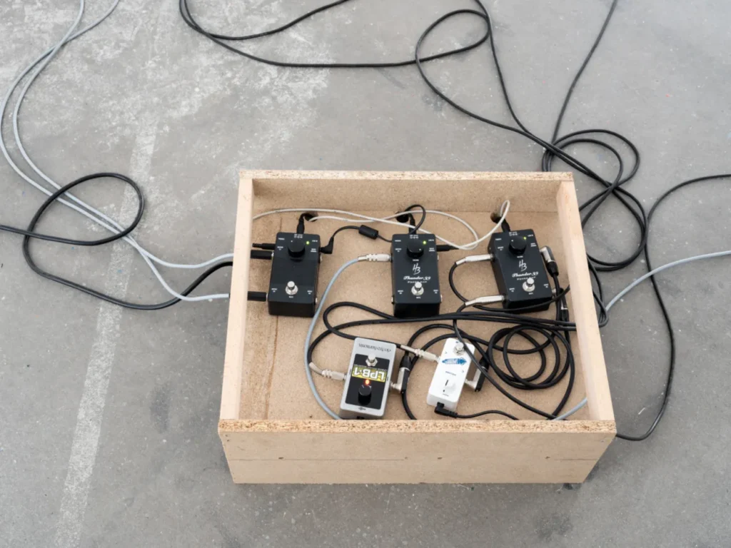 Mehrere schwarze Gitarreneffektpedale und Kabel in einer Holzkiste auf Betonboden, Teil einer Klanginstallation.