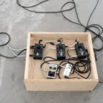 Mehrere schwarze Gitarreneffektpedale und Kabel in einer Holzkiste auf Betonboden, Teil einer Klanginstallation.