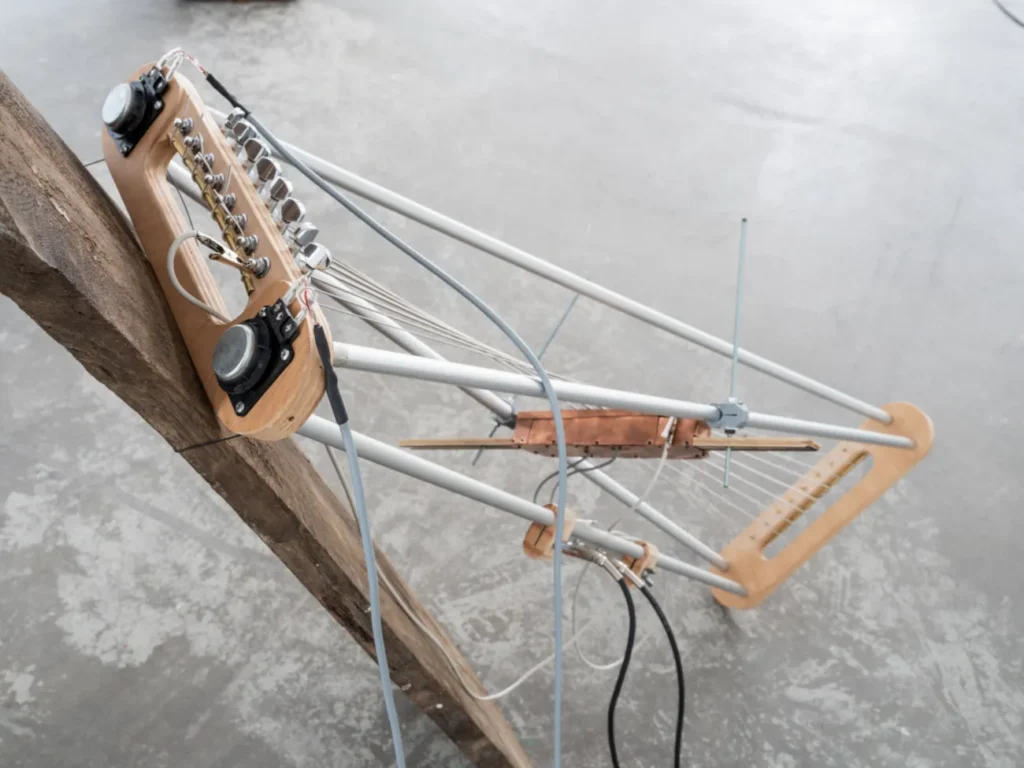 Experimentelles Saiteninstrument aus Holz und Metallrohren, montiert an einem Baumstamm, mit Tonabnehmern und Kabeln.