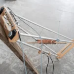Experimentelles Saiteninstrument aus Holz und Metallrohren, montiert an einem Baumstamm, mit Tonabnehmern und Kabeln.