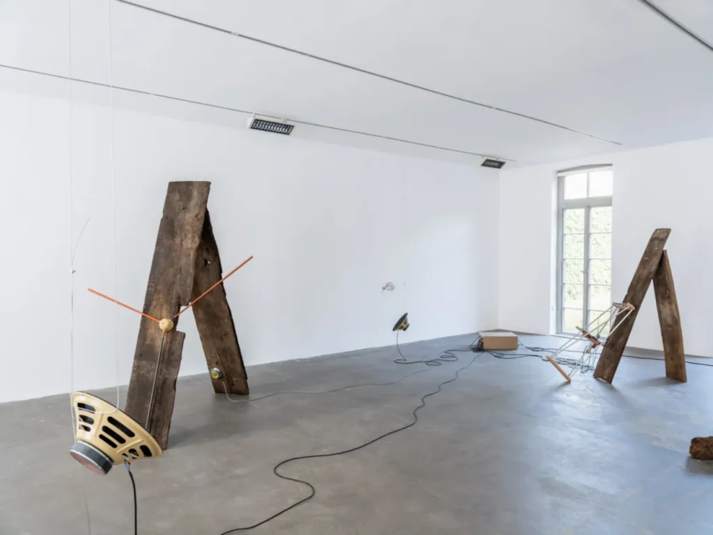 Installationsansicht mit lehnenden Holzbohlen, orangen Stäben, alten Lautsprechern und Kabeln auf grauem Betonboden.