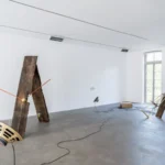 Installationsansicht mit lehnenden Holzbohlen, orangen Stäben, alten Lautsprechern und Kabeln auf grauem Betonboden.